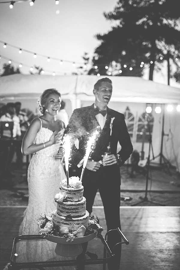 Black-and-white portrait-style shot of a bride and groom standing behind a three-tier naked cake topped with floral decorations and lit sparklers during an outdoor evening reception. The bride wears a fitted strapless lace gown and the groom wears a dark suit with a bow tie; both are laughing and visibly reacting to the sparklers. The cake is displayed on a small glass-topped table. The setting is an outdoor terrace with a white marquee tent and string bistro lights strung overhead, with guests and what appears to be a band setup visible in the background. The image has soft mid-tones with moderate contrast typical of evening ambient light photography.