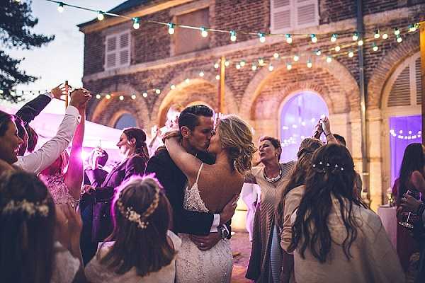 The couple shares a kiss on an outdoor dance floor surrounded by approximately 10-15 guests who are dancing and celebrating around them during an evening reception. The setting is an outdoor courtyard in front of a brick building with arched windows, decorated with string globe lights strung overhead and bathed in warm purple and pink uplighting. The bride wears a low-back lace wedding gown with her blonde hair loosely styled, while the groom is dressed in a dark suit. Guests around them are visibly animated, raising their arms and dancing, with one woman nearby wearing a floral hair accessory. The image is a mid-range portrait shot with a slightly shallow depth of field, giving the surrounding guests a soft blur while keeping the couple in focus.
