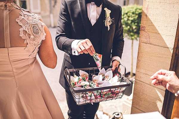 A close-up shot of a groom in a black tuxedo with bow tie and a floral boutonniere reaching into a wire basket filled with paper cones containing colorful flower petal confetti, likely being distributed to guests ahead of a confetti toss. A bridesmaid or wedding party member in a dusty rose/nude dress with lace cap sleeve detail stands to his left, and a guest's hand reaches in from the right to take a cone. The setting appears to be an outdoor or semi-covered area near a stone building, suggesting a French chateau or similar venue. The composition is a medium close-up focusing on the confetti basket detail.