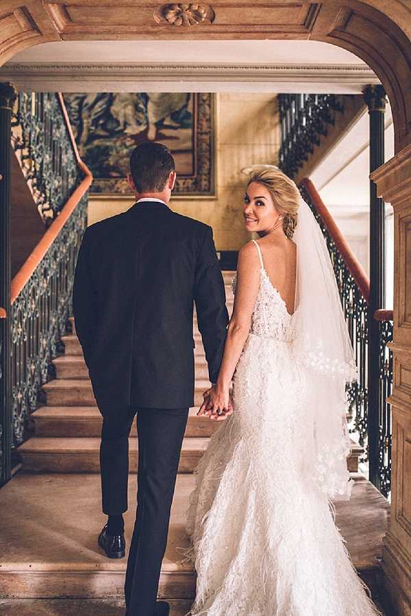A couple walks hand-in-hand up a grand interior staircase, shot from behind at medium distance with the bride glancing back over her shoulder toward the camera. The groom wears a classic black suit, while the bride wears an ivory backless gown with thin spaghetti straps, intricate lace and floral appliqué detailing, and a layered ruffled skirt with feather-trim accents, paired with a cathedral-length veil. The staircase features ornate wrought-iron banisters with a botanical motif, warm wood handrails, stone steps, and a large tapestry or painted mural visible on the landing wall above, suggesting a historic chateau or manor house interior with classic French architecture. Potential venue feature image.