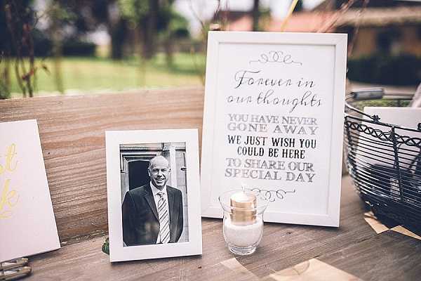 A close-up detail shot of a memorial display set on a wooden table at an outdoor wedding. The display includes a black-and-white photograph of a smiling middle-aged man in a dark suit and tie in a white frame, placed beside a white-framed sign reading 'Forever in our thoughts, you have never gone away, we just wish you could be here to share our special day.' A small gold and glass tealight candle holder sits in front of the sign, and a wire basket is visible on the right edge of the frame. A gold-lettered card is partially visible on the left side of the arrangement.