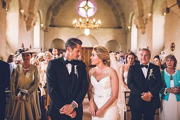 The bride and groom stand together at the altar during a church ceremony, gazing at each other. The groom wears a classic black tuxedo with a bow tie and a small white boutonniere, while the bride wears a fitted spaghetti-strap lace gown with a crystal belt and a sheer veil. The setting is a small historic stone chapel with arched ceilings, a chandelier, wooden pews, and a decorative stained glass rose window above the entrance. Approximately 20-30 guests are visible in the pews behind the couple, including women in formal hats and a man in a dark suit to the right, with warm ambient lighting throughout. Medium-wide shot taken from the aisle looking toward the altar.