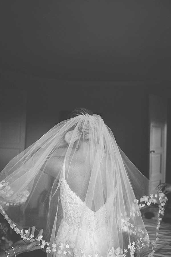 This is a black-and-white getting-ready portrait of a bride indoors, captured as her veil is being placed or adjusted over her face, obscuring her features entirely. The veil features a floral appliqué trim along its edge, and the bride wears a fitted, spaghetti-strap wedding dress with beaded or embroidered detailing on the bodice. A second person's hand and wrist, adorned with a bracelet, is visible at the lower left, suggesting someone is assisting with the veil placement. The image has soft mid-tones with the veil rendered in bright whites against a darker background, shot as a close-up portrait in a room with a visible doorframe.