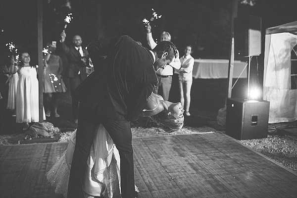 A black-and-white image of a couple's first dance at an outdoor evening reception, captured mid-dip as the groom bends the bride dramatically backward toward the floor on a wooden dance floor. The image has high contrast with deep shadows and bright highlights from artificial lighting on the right side of the frame. Approximately eight to ten guests surround the dance floor holding lit sparklers, with a young girl in a white dress visible on the left. The setting appears to be a partially covered outdoor terrace or pavilion area at night. The composition is a medium wide shot taken from dance floor level, emphasizing the dynamic dip pose.