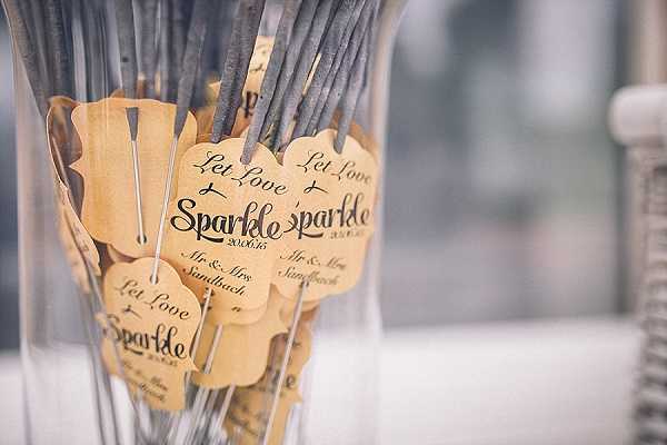 Close-up detail shot of wedding sparklers displayed in a clear glass vase, each tied with a kraft paper tag reading 'Let Love Sparkle' along with a date (20.06.15) and 'Mr. & Mrs. Sandbach' in black calligraphy-style script. The tags have a decorative scalloped edge and are tied to the sparkler wires with grey ribbon. The image has a shallow depth of field with the foreground tags in sharp focus and the background softly blurred, suggesting an indoor reception setting. The styling is classic and personal, using kraft paper and calligraphy as part of a coordinated wedding stationery theme.