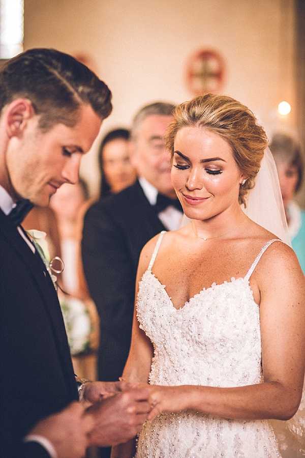 A close-up portrait of a bride and groom during an indoor chapel ceremony, captured at the moment of ring exchange. The groom, wearing a black tuxedo with a bow tie and a white boutonniere, looks down at their joined hands, while the bride smiles softly with her eyes downcast. The bride wears a beaded spaghetti-strap gown with a sweetheart neckline and a sheer veil, with her blonde hair styled in an updo. Several guests are visible in soft focus in the background, seated in the chapel pews. The warm, golden interior lighting and a small cross visible on the back wall confirm a traditional church setting with a classic ceremony style.