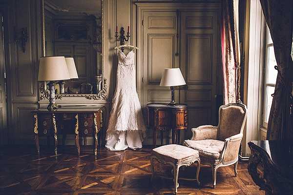 A getting-ready detail shot of an ivory lace wedding dress hanging from a wall-mounted candelabra in an ornate chateau room. The dress features a fitted silhouette with lace overlay and delicate straps. The room is furnished with gilded Louis XVI-style furniture including a marquetry side table with a gold-framed mirror, a matching nightstand with a cream lamp, and an upholstered armchair with footstool in aged ivory fabric, all set on a herringbone parquet floor. The wide interior shot captures warm, dim ambient lighting from table lamps and natural light filtering through a draped window, giving the room a classic French interior aesthetic. Potential venue feature image.