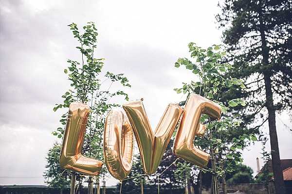 A close-up outdoor detail shot of four gold foil letter balloons spelling 'LOVE,' photographed against an overcast sky. The balloons are rose-gold in tone and are positioned in front of tall leafy branches or small trees used as decorative elements. A stone building is partially visible in the background. The styling suggests a garden or courtyard wedding with a relaxed, modern-rustic aesthetic.