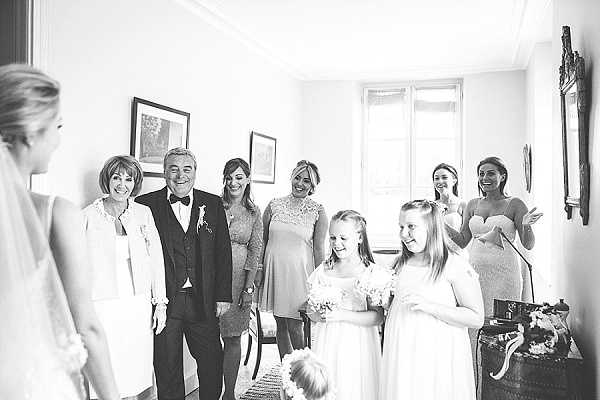 A black-and-white getting-ready moment capturing a first look reveal inside a classic interior room with tall windows and crown molding. The bride, visible from behind on the left in a white gown and veil, faces a group of approximately seven people reacting with smiles and emotional expressions. The group includes an older couple — the man in a dark suit with bow tie and boutonnière, the woman in a light blazer — along with several women in cocktail-length dresses and two young flower girls in white dresses holding small bouquets. The tones are bright and airy with soft contrast. Wide portrait-style shot.