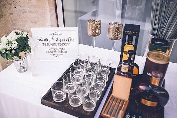 Close-up detail shot of a whiskey and cigar bar station set up at a wedding reception, displayed on a white linen-covered table. A calligraphy-style sign reads 'Whiskey & Cigar Bar – Please Enjoy with compliments of the Bride and Groom' alongside a small silver bucket holding white flowers with green foliage. The bar features multiple whisky tumblers arranged on a dark wooden tray, bottles of Penderyn whisky and at least one other spirit, a box of cigars, tall gold filigree candle holders, and a container of cocktail stirrers or sparklers. The styling is classic and refined with a neutral white and gold palette.