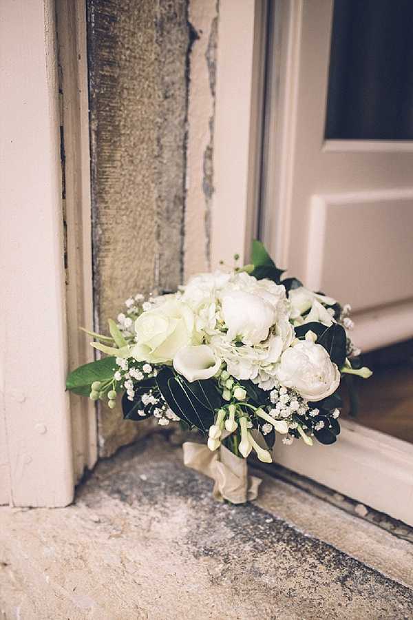 A close-up detail shot of a bridal bouquet resting on a stone threshold against a weathered wooden door frame. The compact, rounded bouquet is composed of white roses, white peonies, white hydrangea, white calla lilies, baby's breath, lily of the valley, and deep green foliage. The stems are wrapped in ivory or cream ribbon. The overall color palette is all-white blooms with lush green accents, giving a classic, understated look.