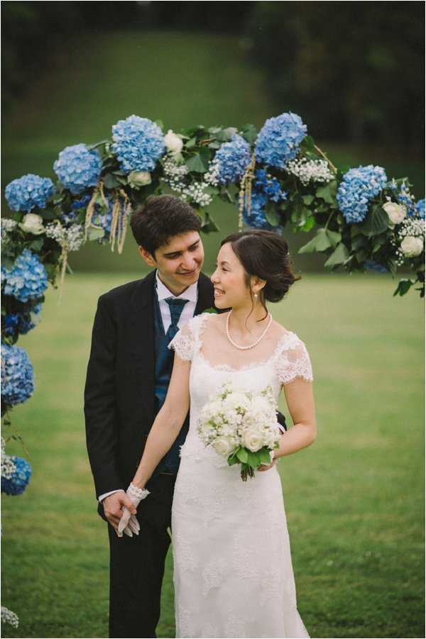 A couple poses together outdoors in front of a circular floral arch decorated with blue hydrangeas, white roses, baby's breath, and green foliage. The bride wears a white lace dress with cap sleeves and an illusion neckline, accessorized with a pearl necklace and wrist corsage, and holds a bouquet of white roses and baby's breath; the groom wears a dark navy suit with a navy tie. The two are looking at each other and smiling in a relaxed portrait composition set on a lawn. The decor palette is blue and white with a classic, garden-party feel.