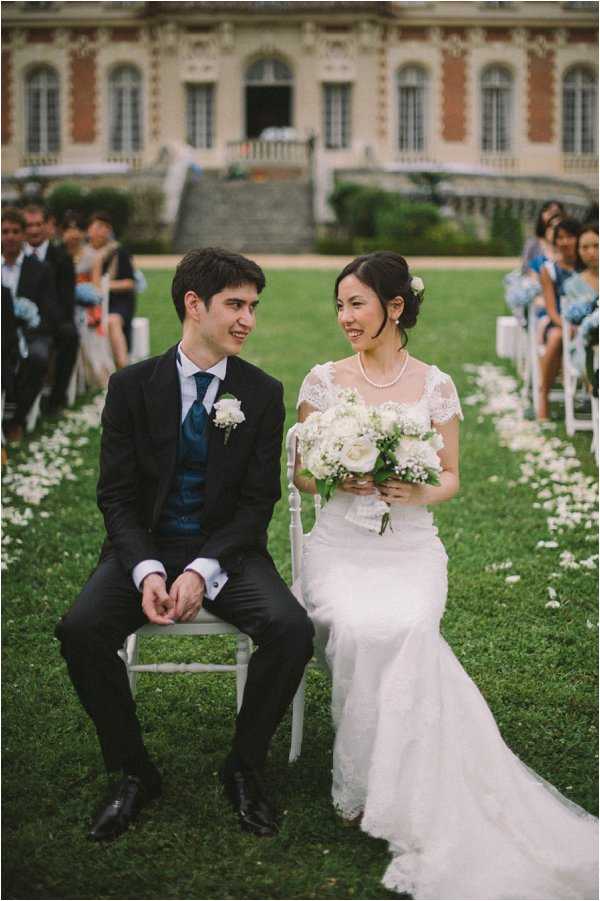 An outdoor wedding ceremony taking place on the lawn of a large French chateau with a red brick and stone facade visible in the background. The couple is seated at the front of the ceremony aisle on white chairs, turning to smile at each other; the groom wears a black suit with a teal/navy waistcoat and tie, and a white boutonniere, while the bride wears a fitted lace gown with cap sleeves and a pearl necklace, holding a bouquet of white roses, peonies, and baby's breath. White flower petals are scattered along the grass aisle, and guests seated in white chairs on both sides are visible in the background, with several women in light blue dresses on the right side. The composition is a medium portrait shot of the couple, with the chateau's grand staircase and balustrade softly out of focus behind them. Potential venue feature image.