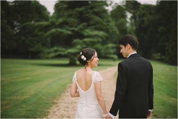 A couple portrait taken outdoors on a gravel path cutting through a wide open lawn, with tall trees in the background. The bride wears a fitted ivory lace gown with a deep V-back, cap sleeves, and button detailing down the spine, with small white floral hair pins in an updo; the groom wears a dark black suit. The two are walking hand in hand away from the camera, with the bride turning slightly to smile over her shoulder. The shot is a medium portrait with soft, overcast natural lighting giving the image a muted, slightly hazy quality.