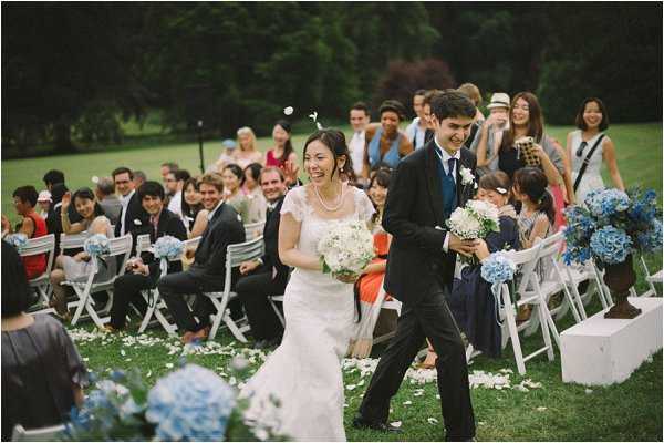 The bride and groom walk back up the aisle following an outdoor ceremony, both smiling broadly. The ceremony takes place on a lawn with white folding chairs arranged in rows on either side of the aisle, which is scattered with white flower petals. Approximately 40-50 guests are seated and standing, many applauding and smiling. The bride wears a white lace gown with short sleeves and a pearl necklace, carrying a round bouquet of white flowers and greenery. The groom wears a black tuxedo with a navy blue tie. The aisle chairs are decorated with arrangements of blue hydrangeas. Wide shot taken from ground level facing the couple as they process toward the camera.