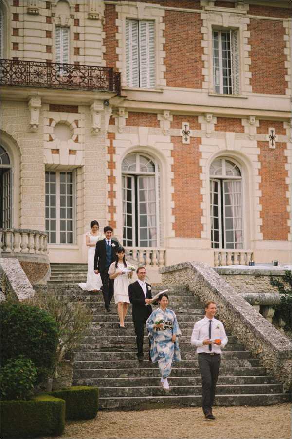 A wedding party descends a wide stone staircase on the exterior grounds of a French chateau, with the couple visible at the top of the stairs — the bride in a white gown and the groom in a dark navy suit with a boutonniere. In front of them, a woman in a white knee-length dress carries a small bouquet, accompanied by a man in a dark suit, while further down the stairs a guest wearing a traditional Japanese blue-and-white kimono walks alongside a man in a white dress shirt and dark trousers. The chateau facade behind them features red brick with cream stone detailing, arched French windows, and a wrought-iron balcony, suggesting a classic French estate setting. The shot is a wide vertical composition capturing the full height of the staircase and chateau facade, with the procession creating a natural diagonal line through the frame. Potential venue feature image.
