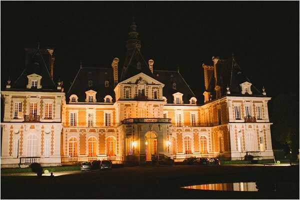 A wide-angle nighttime exterior shot of a large French château fully illuminated by warm amber uplighting against a completely dark sky. The building features classic French classical architecture with mansard roofs, dormer windows, ornate white stone detailing, and a central entrance pavilion with a balcony. No people are visible; the shot focuses entirely on the facade and its reflection in a small pool or water feature in the foreground. Potential venue feature image.