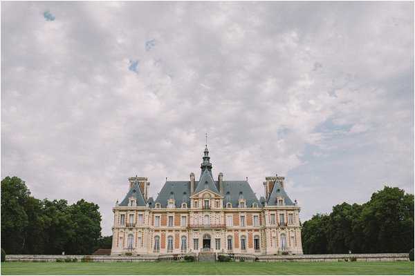Wide-angle exterior shot of a classic French château featuring a symmetrical stone facade with a slate mansard roof, corner pavilions with pointed turrets, and ornate architectural detailing across multiple floors. No people are visible; the image focuses entirely on the building and its formal grounds. The composition is centered and shot from a distance, capturing the full breadth of the château. Potential venue feature image.