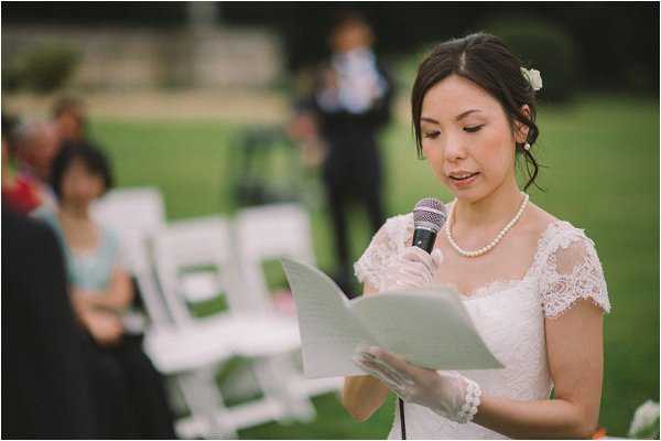 A bride reads her vows from a paper document into a handheld microphone during an outdoor wedding ceremony. She wears a white lace dress with cap sleeves, a pearl necklace, a white lace wrist cuff, and has a small white floral hair accessory pinned into her dark updo. The ceremony is set on a lawn with white folding chairs visible in the background, along with several seated guests and a figure standing further back. The shot is a close-up portrait with a shallow depth of field, leaving the background softly blurred.