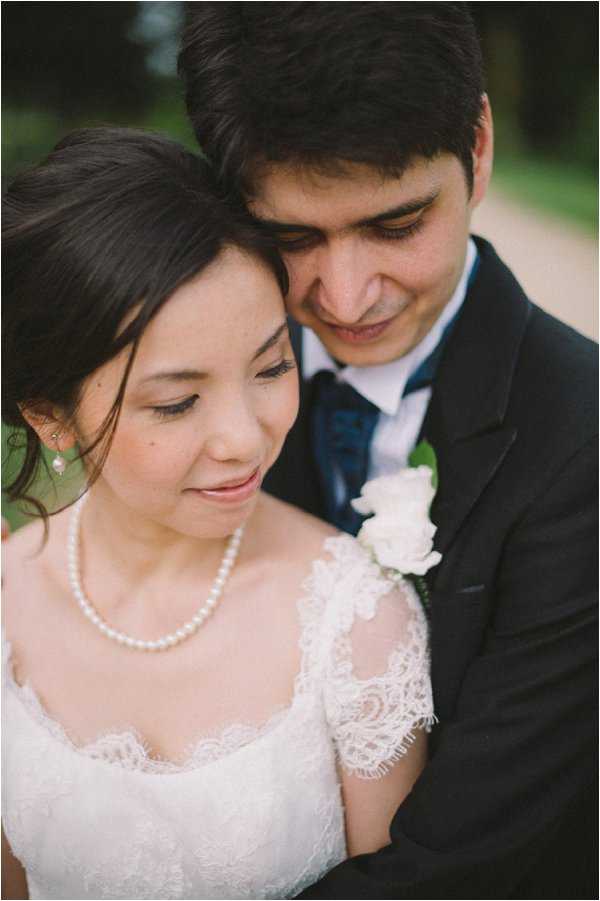 A close-up portrait of a bride and groom outdoors, with a softly blurred green background. The bride wears a white lace dress with short lace sleeves and a scoop neckline, paired with a pearl necklace and small drop earrings; her dark hair is loosely pulled back. The groom wears a dark navy suit with a navy tie and a white rose boutonniere, and he leans his head toward the bride from behind as she looks downward. The shot is tightly framed on the couple's faces and upper bodies, emphasizing their expressions and styling details.