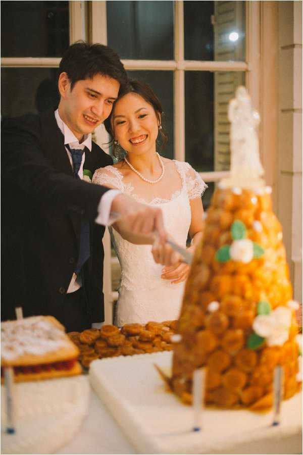 The bride and groom are cutting a traditional French croquembouche at what appears to be an indoor evening reception, photographed in a close-up portrait composition. The croquembouche is a tall cone of choux pastry puffs decorated with white sugar flowers, mint green fondant leaves, and a small bride-and-groom cake topper at the top. The bride wears a white lace dress with cap sleeves and a pearl necklace, while the groom is dressed in a dark navy suit with a blue tie. Additional pastries, including what appears to be a mille-feuille or tart, are visible on the white-clothed table in the foreground.