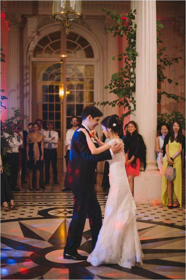 The bride and groom share their first dance in a grand ballroom or orangerie-style interior space, characterized by tall white classical columns, arched French doors, and a striking black-and-white geometric patterned marble floor. The bride wears a fitted white lace gown with an off-the-shoulder neckline, and the groom wears a dark navy suit with a floral boutonnière. Approximately 15–20 guests form a circle around the couple, watching the dance; notable guest attire includes a bright yellow maxi dress and a coral mini dress. The space is lit with warm amber overhead lighting and red-pink uplighting on the walls, creating a warm evening atmosphere, with trailing green vines decorating one of the columns. The shot is taken from a medium distance, capturing both the couple in full length and the surrounding guests.