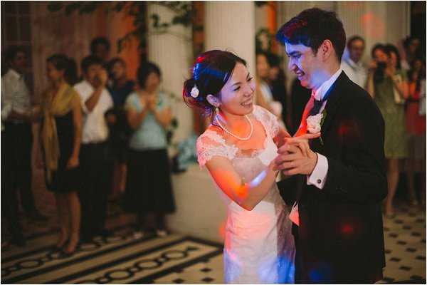 The bride and groom share their first dance inside a formal venue with classical columns and a black-and-white patterned tile floor visible in the background. The bride wears a short-sleeved lace dress in an off-white or blush tone, a pearl necklace, and a floral hair accessory, while the groom is dressed in a dark suit with a white boutonniere. Warm orange and blue stage lighting illuminates the dance floor, creating a colorful atmosphere. Approximately 20-30 guests stand and watch in the background, and the image is a medium-wide portrait shot with the couple sharp in the foreground and guests softly blurred behind them.
