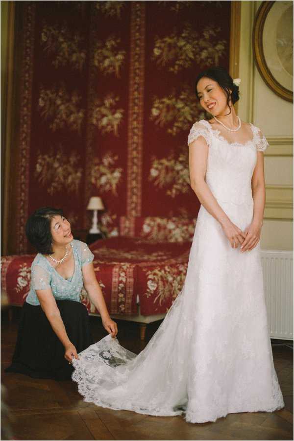 A getting-ready moment captured indoors in what appears to be a chateau or manor house bedroom, featuring ornate red floral wall panels and gold-framed artwork in the background. The bride stands in a white lace cap-sleeve gown with a lace train, wearing a pearl necklace and a white floral hair accessory, smiling down at a woman kneeling beside her who is adjusting and arranging the lace train. The second woman, likely the bride's mother, wears a light blue sequined top and black skirt with a pearl necklace. The composition is a medium portrait shot with warm, slightly vintage toning, taken on dark hardwood floors.
