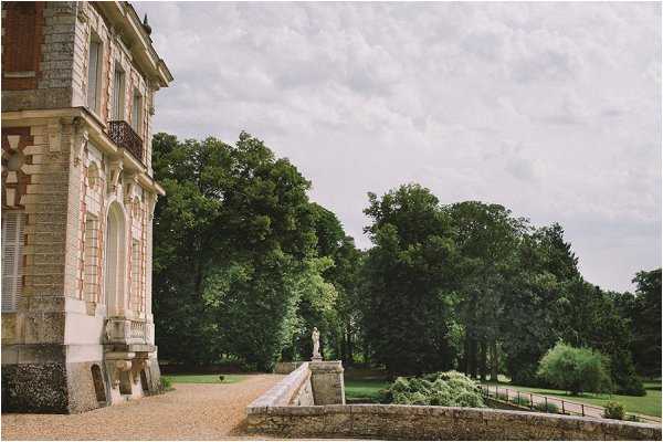 Wide exterior shot of a French chateau venue, showing the ornate facade with arched windows, brick and stone detailing, and a wrought-iron balcony along the left side of the frame. A gravel pathway extends along the building leading to a balustrade terrace featuring a classical stone statue. No people are visible in this image. Potential venue feature image.