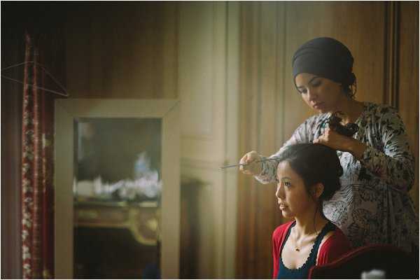A getting-ready scene showing a hair stylist wearing a dark head wrap and a floral-print top working on a seated woman's updo hairstyle, using what appears to be bobby pins or hair pins. The seated woman is dressed in a red and navy top. The setting appears to be an indoor room with warm, dim ambient lighting, sheer curtains diffusing natural light from a window, and an ornate mirror reflecting a decorative fireplace mantel in the background, suggesting a chateau or historic venue interior. The image is a medium portrait shot with a shallow depth of field and warm, slightly dark tones.