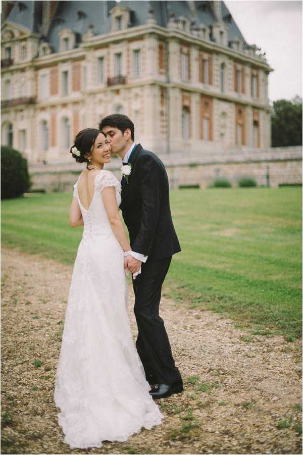 A couple portrait taken outdoors on the gravel grounds of a large French chateau with pale stone and brick facade and a mansard roof. The groom, wearing a dark navy suit with a white boutonniere, kisses the bride on the cheek while she turns to smile at the camera. The bride wears a white lace gown with cap sleeves and a low open back, and has her dark hair pinned up with small white floral hair accessories. They are holding hands, with the full skirt and short train of her dress visible. The composition is a medium full-length portrait shot with the chateau softly blurred in the background. Potential venue feature image.