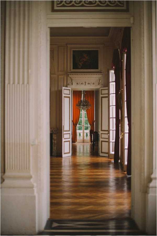 An empty interior corridor of a grand chateau or palace-style venue, shot as a wide perspective through a series of doorways leading into successive rooms. The architecture features tall fluted columns, ornate white-painted wood paneling with gold detailing, decorative plasterwork moldings, and a classical oil painting mounted above a fireplace mantel. The floors are polished herringbone parquet wood in warm honey and amber tones. Deep burnt-orange/rust-colored floor-length curtains frame the tall windows and French doors at the far end, through which greenery is faintly visible. A small crystal chandelier hangs in the middle room. No people are present. The composition is a straight-on perspective shot emphasizing depth and the enfilade room layout. Potential venue feature image.
