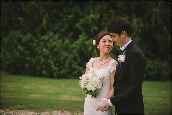 A couple portrait taken outdoors on a lawn with tall dark green hedges in the background. The bride wears a fitted lace dress with cap sleeves and holds a bouquet of white hydrangeas and white roses with green foliage; she also wears a pearl necklace and a small white floral hair accessory. The groom is dressed in a dark navy suit with a white boutonniere and leans in close toward the bride, who smiles at the camera. The portrait is shot at medium range with a shallow depth of field, keeping focus on the couple while the background is softly blurred.