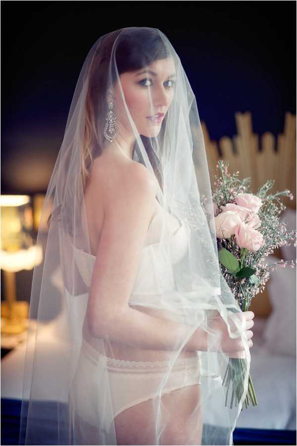 A bridal boudoir portrait shot indoors, likely in a hotel room or bedroom setting with a lamp and decorative headboard visible in the background. The bride wears white lingerie including a lace garter and a sheer, lace-edged cathedral-length veil draped over her head and body, paired with large chandelier crystal drop earrings. She holds a small bouquet of blush pink roses and pink waxflower accents tied with a white ribbon. The image is a close-to-medium portrait composition with soft, warm ambient lighting, and the overall styling leans toward a classic bridal boudoir aesthetic.