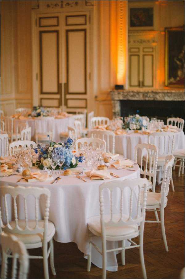 A wedding reception setup inside a classic French chateau ballroom featuring multiple round tables dressed in white floor-length linens, surrounded by white Napoleon-style chairs with cream cushions. The foreground table is set with crystal wine glasses, peach folded napkins, bread rolls, and a low floral centerpiece of blue hydrangeas, delphinium, and cream roses. Warm amber uplighting illuminates the wood-paneled walls and a marble fireplace is visible in the background. The decor palette combines white, peach, and cornflower blue against the room's parquet flooring and ornate architectural details. Wide shot taken from a slightly elevated angle showing three or four guest tables in the reception space. Potential venue feature image.