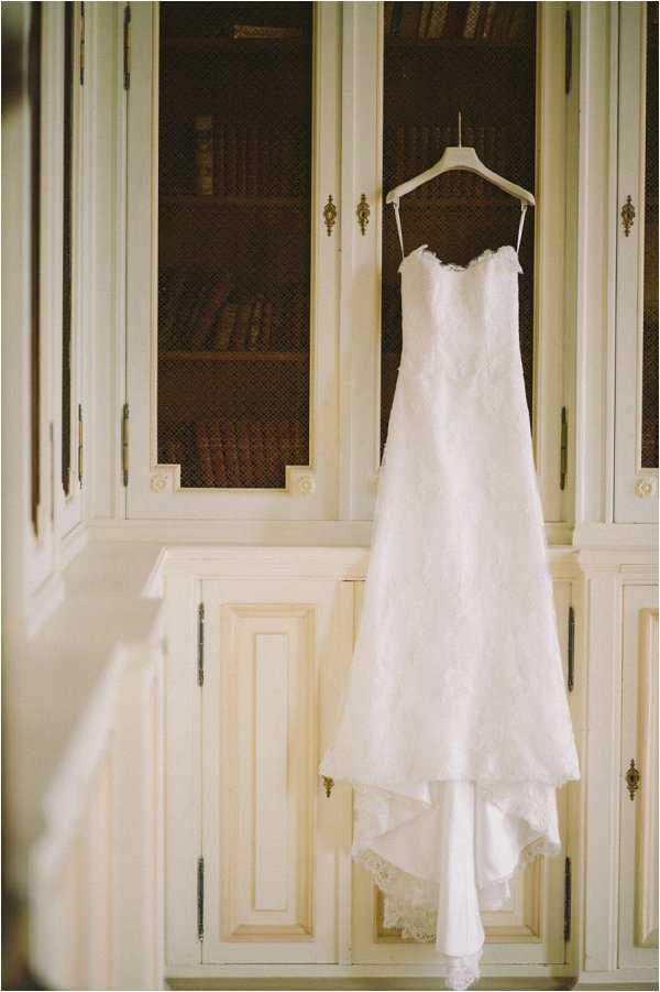 A getting-ready detail shot of an ivory lace wedding gown hanging on a white hanger in front of a built-in bookcase with glass-fronted cabinet doors painted in a cream finish with brass hardware. The dress is a fitted, strapless A-line silhouette with all-over lace overlay, thin spaghetti straps, a sweetheart neckline, and a lace-trimmed hem. The bookcase behind the dress contains rows of antique-looking leather-bound books visible through the glass panels, suggesting an interior room of a chateau or manor house. The image is a medium vertical shot with soft, warm ambient lighting.