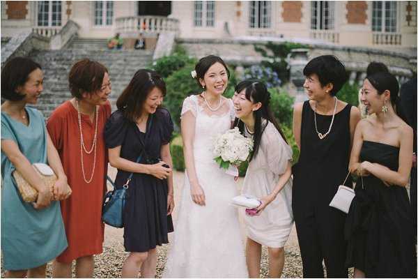 A bride stands outdoors with a group of six female guests in a candid, laughing moment, likely during a cocktail hour or post-ceremony gathering. The setting is the gravel grounds in front of a classical French chateau or manor, with stone balustrades and steps visible in the background. The bride wears a white lace-cap-sleeve gown with a pearl necklace and holds a compact bouquet of white roses and greenery; the guests wear a mix of casual cocktail attire in teal, rust-red, navy, cream, and black. The shot is a mid-length portrait capturing genuine expressions of joy among the group.