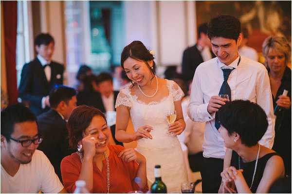 The bride and groom are mingling with seated guests during the wedding reception, likely in a ballroom or grand interior room with warm ambient lighting. The bride wears a short white lace-sleeved dress with a pearl necklace and holds a champagne flute, while the groom is in a white dress shirt with a navy tie and holds a wine glass. The guests, approximately 10-12 visible, are dressed in formal attire including a woman in a rust-orange dress with a beaded necklace seated in the foreground. The image is a candid mid-shot capturing a relaxed, convivial moment with natural smiling expressions, shot with a warm color tone and shallow depth of field that blurs the background guests.