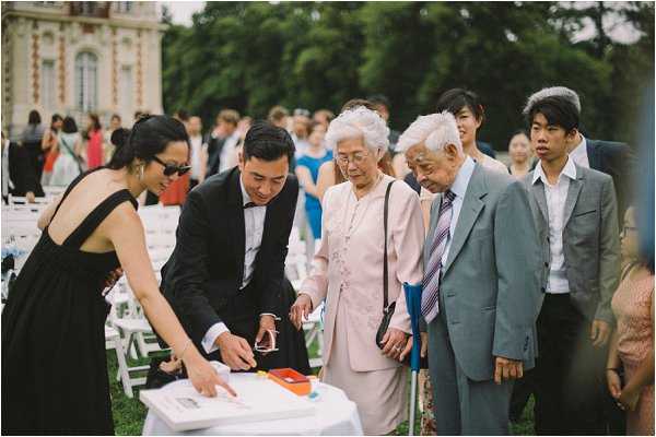 An outdoor wedding scene on a lawn where a man in a black suit is directing two elderly guests — a woman in a pale pink dress and a man in a grey suit with a striped tie — toward a guest book or signing table with colorful ink pads, assisted by a woman in a black halter dress wearing sunglasses. Rows of white folding chairs are visible in the background, along with a large group of guests and the ornate white facade of what appears to be a French chateau. The setting and guest count suggest a sizeable formal outdoor ceremony. This is a candid mid-shot capturing the guest arrival and signing activity.
