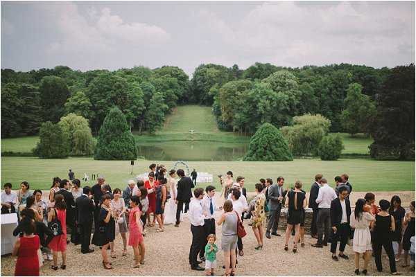 A wide-shot overhead view of a wedding cocktail hour taking place on a gravel terrace, with approximately 40-50 guests mingling in small groups. Guests are dressed in summer attire including coral, red, and navy outfits, with the bride visible in a white gown near the center of the crowd. In the background, a circular floral arch ceremony structure remains set up on an open lawn leading down to a long rectangular reflecting pool or canal flanked by rows of tall trees, creating a formal French garden perspective. The setting appears to be a classic French chateau estate with formal landscaped grounds. Potential venue feature image.