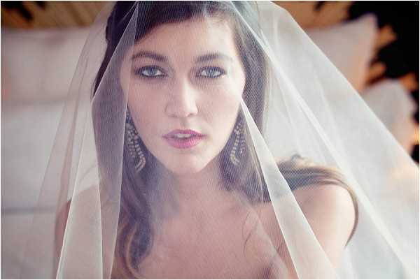Close-up portrait of a bride draped in a sheer ivory veil that falls over her face and shoulders. She has dark hair styled up, blue-grey eyes with defined eye makeup, and a soft pink lip. She wears large chandelier earrings with an intricate silver and crystal design. The background is soft and out of focus, suggesting an indoor setting with warm, diffused natural light. The composition is an intimate, slightly hazy close-up with a soft-focus, film-like quality.