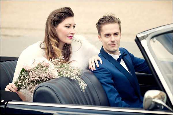A couple sits together in the back seat of a vintage black convertible car in what appears to be a portrait or transportation shot. The bride wears a white or cream wrap/jacket and holds a loose, garden-style bouquet of blush pink blooms and delicate dusty pink waxflower sprigs. The groom wears a navy blue suit with a white dress shirt and black bow tie, with his arm resting along the back of the seat. The styling has a retro, mid-century feel consistent with the classic car. The image is a close-up portrait composition shot from outside the car, with a neutral, blurred background.