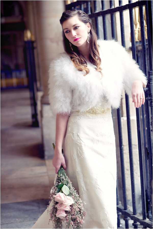 A bridal portrait shot outdoors in an urban architectural setting, featuring a woman in an ivory lace wedding gown with a beaded waistband, wearing a white feather bolero jacket over it. She holds a loosely gathered bouquet of blush pink roses and dried lavender-toned foliage at her side, and wears chandelier earrings with bold fuchsia lip color, giving the look a vintage-glamour styling. Her dark hair is worn in soft waves with a side-swept style. The composition is a medium close-up portrait taken alongside black iron railings with stone columns visible in the soft-focus background.