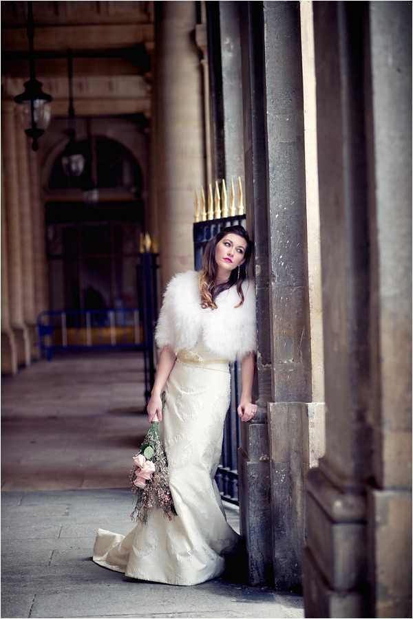 A bridal portrait shot outdoors beneath a classical stone arcade, likely in Paris, featuring a bride standing between large stone columns. The bride wears a fitted ivory brocade gown with a train and a white feather bolero shrug, paired with a gold sash at the waist and a gold spiked crown headpiece. She holds a loosely gathered bouquet of blush pink roses and dried botanicals at her side. Her styling is theatrical and fashion-forward, with bold pink lip color and long wavy dark hair. The composition is a full-length portrait with shallow depth of field, blurring the arched colonnaded walkway and ornate black-and-gold iron gates in the background.