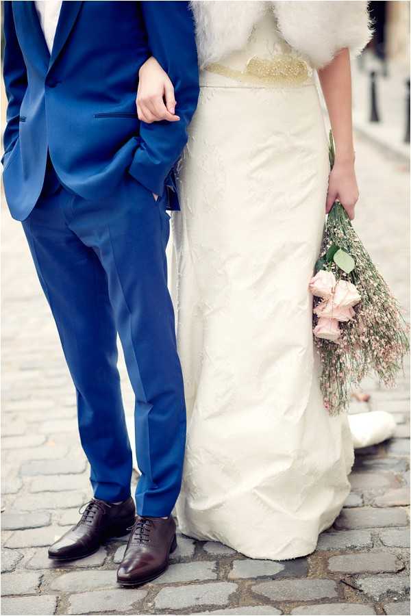A close-up detail shot of a bride and groom standing together on a cobblestone street, cropped from the waist down. The groom wears a cobalt blue suit with slim-cut trousers and brown leather oxford shoes. The bride wears an ivory textured gown with a gold embellished waistband and a white fur stole or wrap visible at the top of the frame. She holds a loosely arranged bouquet featuring blush pink roses and dried pampas-style grasses with eucalyptus foliage, giving the arrangement a soft, romantic feel. The styling combines a classic silhouette with a cool-toned color palette of cobalt and ivory accented by blush florals.