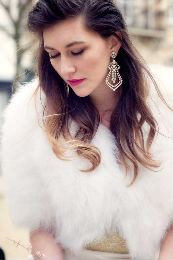 Close-up portrait of a bride outdoors in an urban setting, with blurred building architecture visible in the background. She wears a white faux-fur or feather wrap stole over what appears to be a gold sequined dress, with large Art Deco-style chandelier earrings featuring crystal or rhinestone detailing. Her hair is styled in loose waves with an ombre effect from dark brown to honey blonde, worn half-up. Her makeup features a bold fuchsia-pink lip. The image has a soft, bright, warm tone with shallow depth of field.