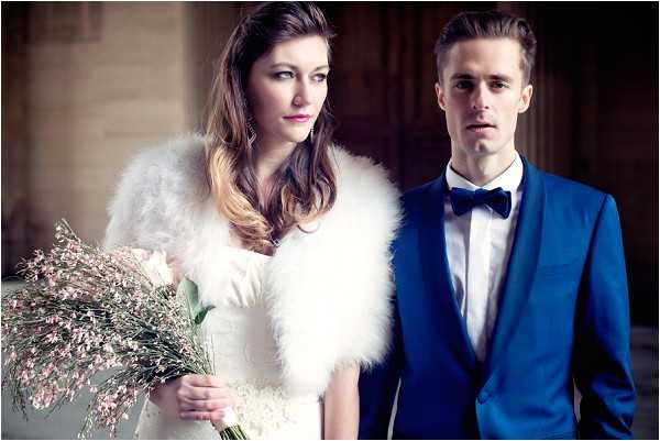 A couple portrait shot in a close-up composition, set indoors against what appears to be a stone archway or classical interior. The bride wears a fitted white dress with a white feather or faux-fur stole and holds a loose, wildflower-style bouquet featuring blush pink blooms and wispy dried grasses. The groom wears a cobalt blue suit with a white shirt and a black bow tie. Both subjects face slightly different directions rather than the camera, giving the image a editorial, fashion-forward feel. The styling is modern and polished with a cool-toned color palette of white, cobalt blue, and soft blush.