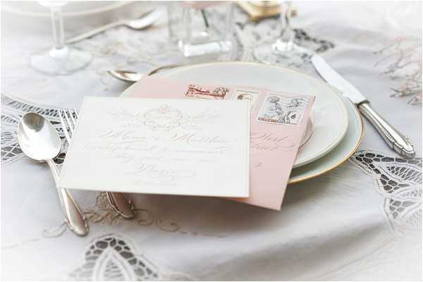 A close-up detail shot of a wedding reception place setting featuring a white calligraphy menu card and a blush pink envelope with vintage-style postage stamps resting on a stacked white and gold-rimmed charger plate. The table is dressed with a white lace-embroidered tablecloth, and silver flatware is arranged on either side of the plate. Crystal glassware is partially visible in the background. The overall decor palette is soft blush, ivory, and gold, consistent with a classic, romantic styling approach.