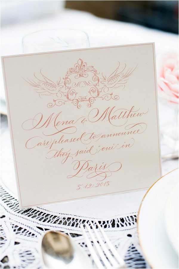 Close-up detail shot of a wedding announcement card propped on a table, reading 'Mena & Matthew are pleased to announce they said oui in Paris, 5.12.2015' in rose gold calligraphy script on an ivory background, topped with an ornate crest illustration featuring swallows and scrollwork in matching rose gold ink. The card is displayed on a white crocheted lace table linen alongside a silver spoon and a gold-rimmed white plate, with a blush pink rose and a glass partially visible in the background. The stationery design follows a classic French-inspired style with formal script lettering. The overall table palette is ivory, white, and blush pink.