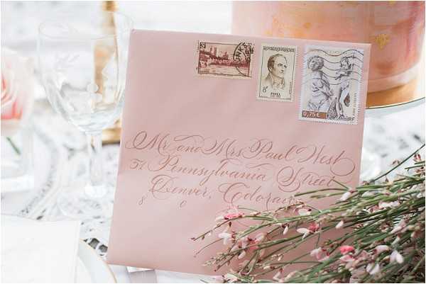 Close-up detail shot of a blush pink envelope addressed in white calligraphy script to 'Mr. and Mrs. Paul West, 57 Pennsylvania Street, Denver, Colorado,' decorated with three vintage postage stamps including what appears to be a French 0.75€ stamp. The envelope is propped against a tiered wedding cake with a watercolor-style peach and gold design, partially visible in the background. Small pink wax flower sprigs are arranged in the foreground, and crystal glassware is softly blurred in the background left. The overall styling palette is blush pink, gold, and ivory with a romantic classic aesthetic.